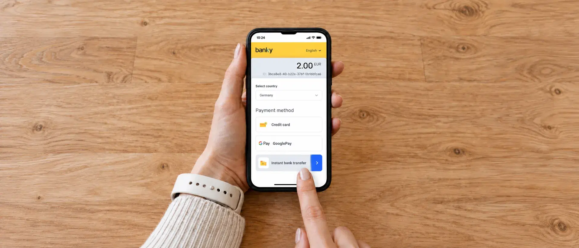 Two hands holding a phone flat over a pale wooden table, seen from above, the Banky checkout open on it and a finger about to press instant bank transfer in the list of payment methods.